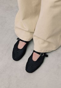 Zwarte gevlochten platte schoenen met een open teen, voorzien van een kleine enkelband met een zilveren gesp. Draag ze met losse beige broeken.