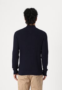 Marineblauer Strickpullover mit strukturiertem Muster, gerippten Bündchen und Kragen. Der Stoff scheint weich zu sein, mit einem geraden Saum. Über beigen Hosen getragen.