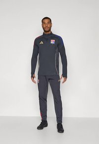 Muž stojí, oblečený v tmavosivom športovom tréningovom obleku s červenými a modrými pruhmi a logami futbalového klubu Lyon.