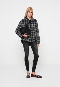 Cămașă supradimensionată în carouri alb-negru, top negru, blugi skinny negri și pantofi din piele patentat burgund; ținând o geantă neagră. Fundal alb simplu.