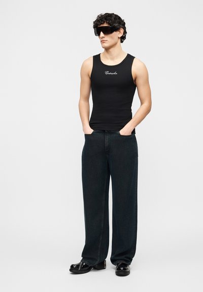 Jeune homme aux cheveux bouclés portant de grandes lunettes de soleil noires, un débardeur noir ajusté, un jean sombre ample et des chaussures noires brillantes.