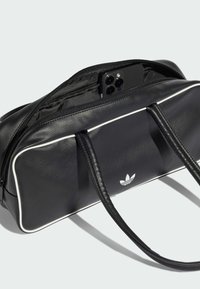 Czarna skórzana torba na ramię z białym obszyciem. Posiada podwójne uchwyty, zapięcie na zamek i logo Adidas z przodu. Wnętrze pokazuje telefon.