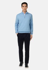 Un pull bleu clair à col zippé avec un ourlet côtelé, associé à un pantalon en velours côtelé noir et des baskets noires, porté par une silhouette masculine se tenant debout.