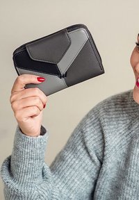 Gray and black wallet with geometric design, smooth texture, and zip closure, held in a hand with red nail polish against a neutral background.
