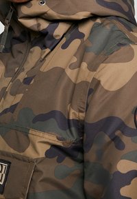 Chaqueta de camuflaje en tonos tierra (marrón, verde, negro) con acabado resistente al agua, que cuenta con un bolsillo frontal y capucha con cordón.