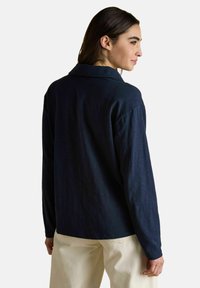 Tumeate juustega naine kannab tumesinist pika varrukaga särk ja kreemikat pükste komplekti, seistes küljega seina vastu taustal, mis on ühtlane.