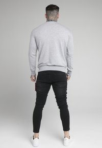 Sudadera gris con cuello de canalé, jeans ajustados negros, zapatillas blancas. Detalle de tatuaje visible en el cuello, accesorio rojo sujeto a los jeans.