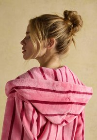 Różowy szlafrok w paski z kapturem, wykonany z pluszowego materiału. Posiada wyraźne ciemniejsze pasy oraz miękką fakturę. Brak widocznych dodatkowych detali.