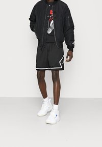 Chaqueta bomber negra, camiseta gráfica negra con acentos rojos, pantalones cortos de malla negros con ribetes blancos y zapatillas blancas con detalles azules.