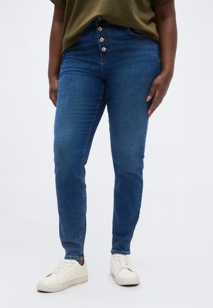 Persona con vaqueros pitillo azules de talle alto con cuatro botones y zapatillas blancas, de pie frente a un fondo blanco liso.