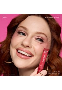 Femme souriante aux cheveux rouges tenant un gloss à lèvres NYX Jelly Job rouge près du visage, portant un fard à paupières rose et des lèvres roses brillantes, fond rose.