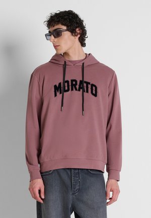 Junger Mann, der einen mauvefarbenen Hoodie mit schwarzem "MORATO"-Text und schwarze Sonnenbrille trägt, vor einem schlichten hellgrauen Hintergrund steht.