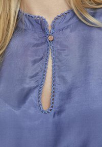 Lila blus med vågformad halsringning, en nyckelhålsöppning och en dekorativ knapp i mitten. Slät tyg med en lätt glans.