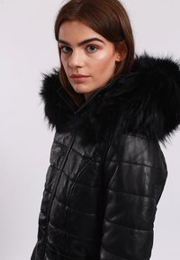 Svart läderjacka med kviltad design, försedd med en voluminös huva av svart konstpäls. Dragkedjestängning och enhetlig färg genom hela jackan.