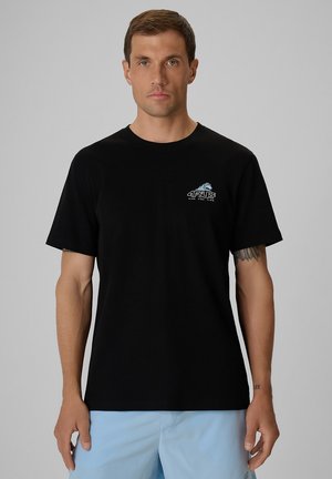 Mężczyzna zwrócony przodem, ubrany w czarną koszulkę z krótkim rękawem z małym białym i niebieskim logo "4F Surf" na piersi oraz jasnoniebieskie szorty.