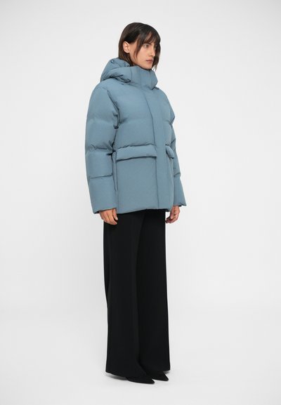 Svetlo modra puhovka s kapuco, ki ima kvadratno silhueto, dva sprednja žepa in teksturiran videz, v kombinaciji z črnimi širokimi hlačami.