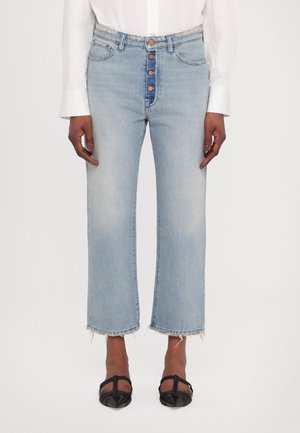 Donna che indossa jeans a vita alta blu chiaro con orli sfilacciati, bottoni in rame, una camicia bianca a maniche lunghe e scarpe nere a punta con cinturini.