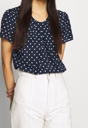 Vrouw die een marineblauwe blouse met korte mouwen en witte stippen draagt en een witte broek met hoge taille, met de armen achter de rug.