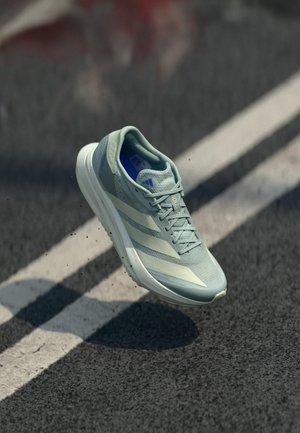 Vaaleansininen Adidas-juoksukenkä ilmassa asfalttitien yllä, jossa valkoiset kaistamerkinnät ja pieniä tien pinnasta irronneita roskia.