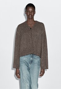 Brązowy, prążkowany krótki sweter z dekoltem w serek i zapięciem na guziki. Noszony na jasnoniebieskich dżinsach. Materiał wydaje się miękki i dzianinowy.