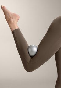 Hnědé elastické legíny s kruhovým, metalicky stříbrným objektem umístěným těsně v ohybu lokte. Hladký povrch a přiléhavý design.