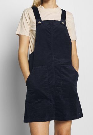 Vrouw draagt een beige T-shirt en een marineblauwe ribfluwelen tuinbroekjurk met voorzakken en verstelbare schouderbanden.