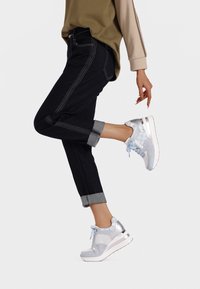Sneakers con una toma superiore in rete blu chiaro e argento, caratterizzati da una suola bianca, accenti decorativi e una linguetta testurizzata. Indossati con jeans neri risvoltati.