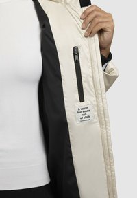 Jakke med lynlås i creme og sort farveblokke, med et stofmærke der står "Et varmt kram lavet af stof," blød tekstur.