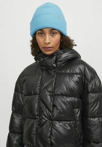 Czarna kurtka puchowa o połyskliwej powierzchni, z wysokim kołnierzem i czarnymi zatrzaskami. Połączona z jasnoniebieską, prążkowaną czapką.