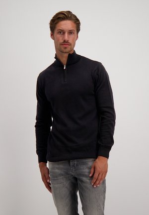 Zwarte zip-up trui met een hoge kraag, gemaakt van zachte stof. Lange mouwen en een ontspannen pasvorm, gecombineerd met lichtgrijze jeans.