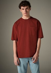 Tricou cu mâneci scurte, culoare ruginie, cu guler rotund și buzunar pe partea stângă a pieptului, confecționat dintr-un material texturat. Purtat cu o pereche de blugi deschis la culoare.