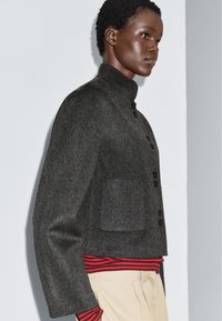 Kobieta z krótkimi, kręconymi włosami, ubrana w ciemnoszary wełniany żakiet zapinany na guziki, pod którym ma na sobie czerwono-czarną pasiastą bluzkę oraz beżowe spodnie, zwrócona w prawo.