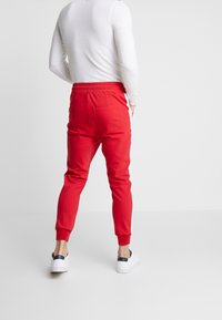 Mann trägt rote Jogginghosen mit weißen Seitenstreifen, ein weißes Langarmshirt und weiße Turnschuhe und steht mit den Händen in den Taschen.