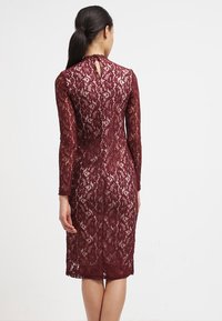 Robe en dentelle bordeaux avec manches longues, silhouette ajustée et détail goutte d'eau dans le dos. Motif floral texturé sur toute la surface.