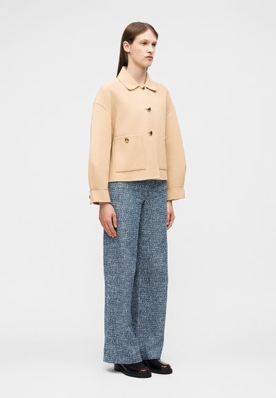 Γυναίκα στέκεται φορώντας ένα μπεζ σακάκι με κουμπιά και μπροστινές τσέπες, μπλε υφασμάτινο παντελόνι με φαρδιές φάσες και μαύρα παπούτσια, σε απλό φωτεινό φόντο.
