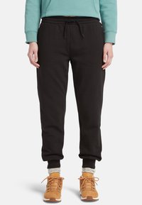 Pantalon de survêtement en polaire noire avec une taille élastique et un cordon de serrage, s'évasant vers la cheville; associé à des chaussures beiges et des chaussettes grises.