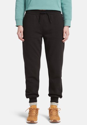 Pantalon de survêtement en polaire noire avec une taille élastique et un cordon de serrage, s'évasant vers la cheville; associé à des chaussures beiges et des chaussettes grises.