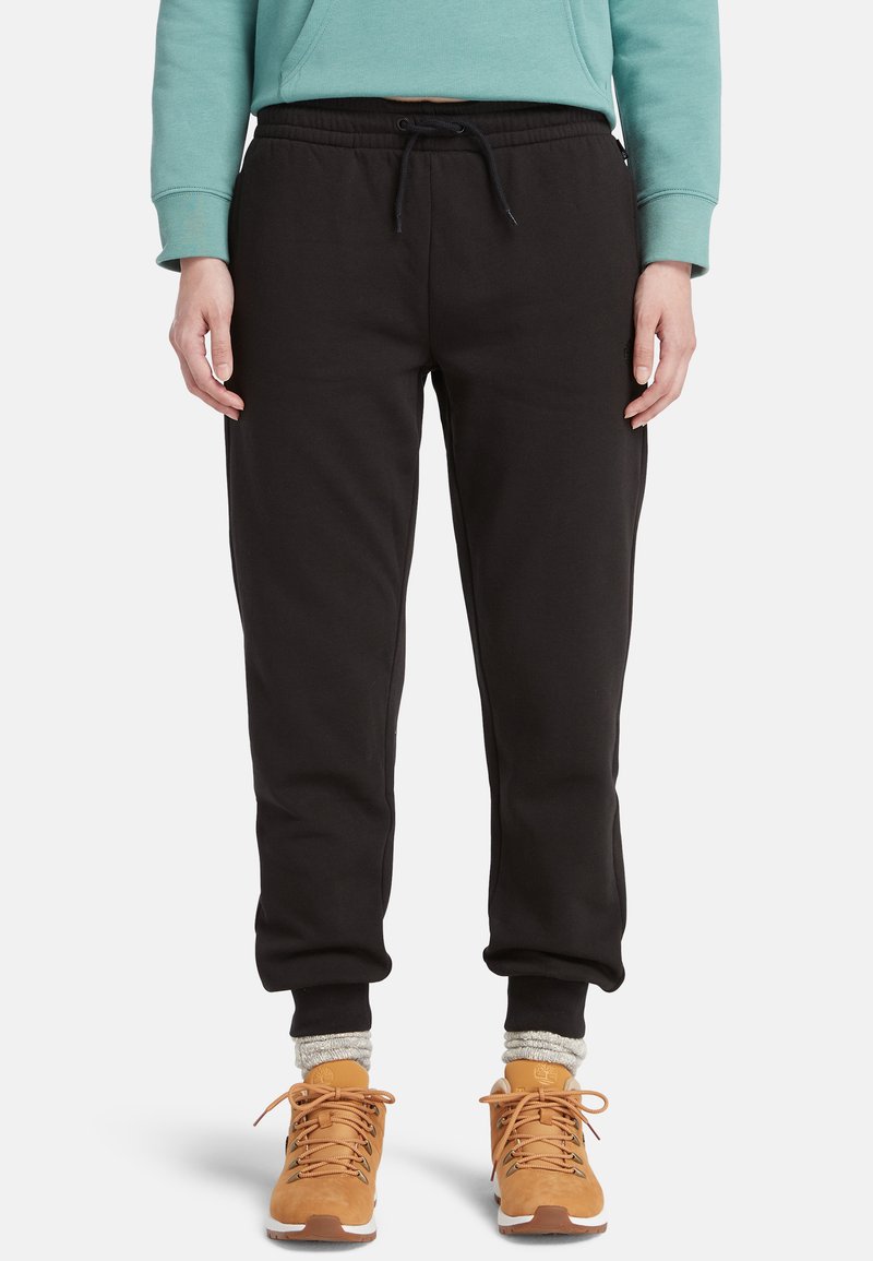 Black fleece sweatpants with an elastic waistband and drawstring, tapering towards the ankle; paired with tan shoes and gray socks.