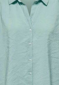 Chemise bleu clair à boutons, en tissu texturé, avec un col et cinq boutons blancs sur le devant.