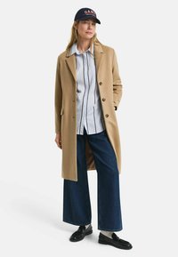 Beige wool coat, buttoned, over a blue and white striped shirt, paired with wide-leg denim pants and black loafers, topped with a navy cap.