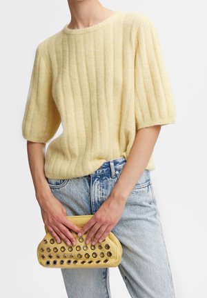 Person som bär en gul ribbad kortärmad tröja och ljusblå jeans, håller en gul kuvertväska med metallöglor.