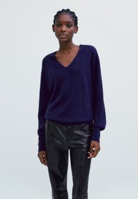 Pull en V bleu marine avec des manches longues légèrement bouffantes, porté avec un pantalon en cuir noir. Texture douce, coupe décontractée et design minimaliste.