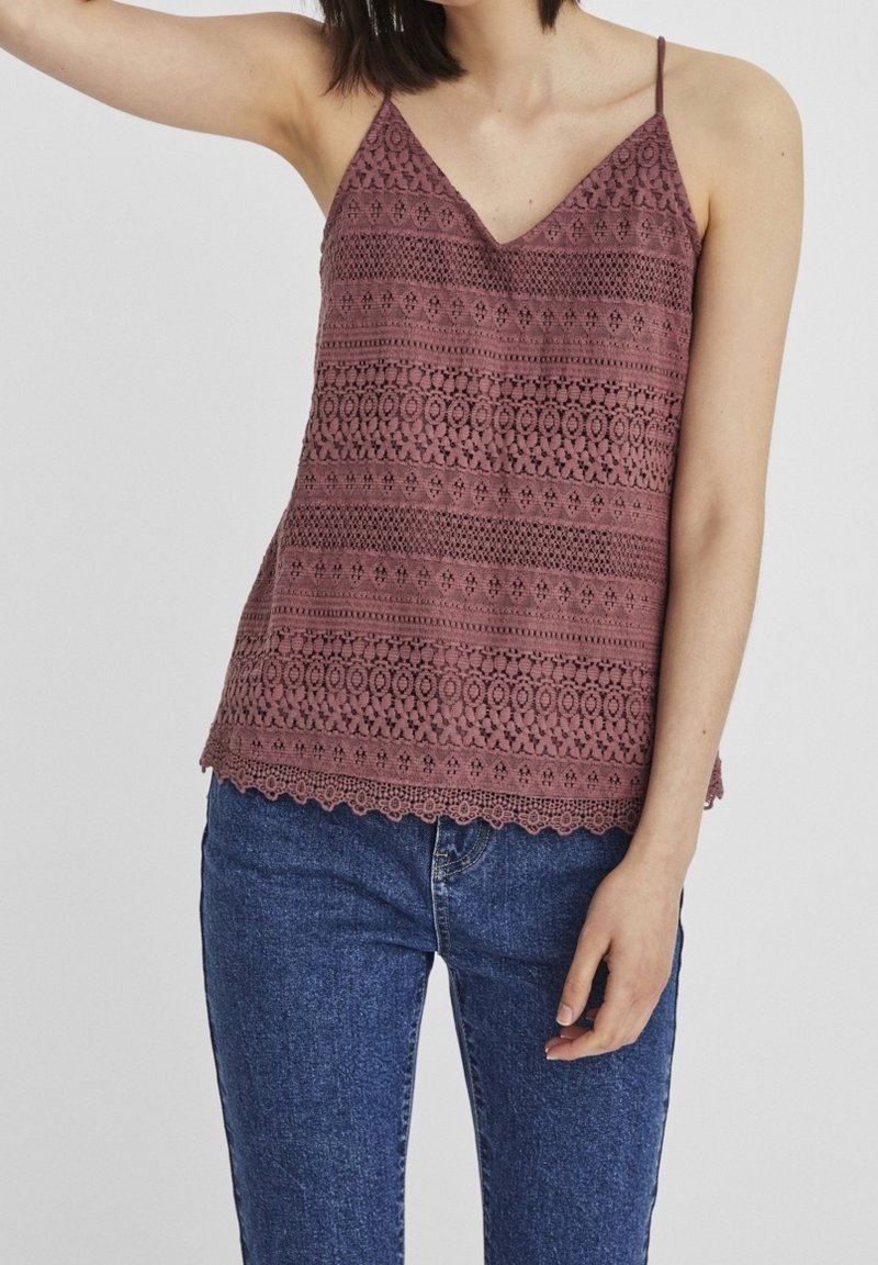 Femme portant un haut en dentelle bordeaux sans manches avec des fines bretelles et un jean en denim bleu sur un fond uni.