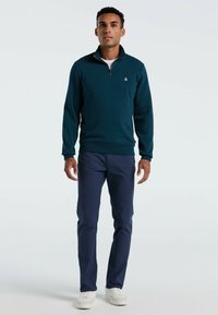 Man staat in een teal kwart-rits trui, marineblauwe broek en witte sneakers tegen een eenvoudige lichte achtergrond.