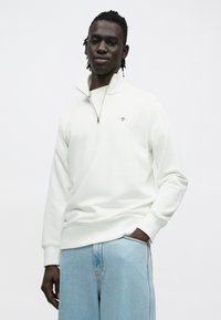 Fehér cipzáros pulóver bordázott mandzsettával és szegéllyel, kis hímzett logóval. Világoskék farmernadrággal párosítva.