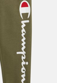 Pantalons de survêtement vert olive avec un grand logo "Champion" blanc sur le côté et un accent rouge dans le logo. Tissu doux avec une texture lisse.