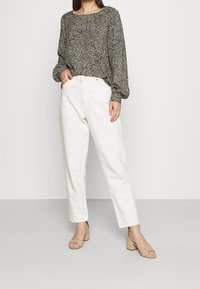 Bluse mit Leopardenmuster in Schwarz und Beige mit Ballonärmeln, kombiniert mit hochgeschnittenen, cremefarbenen Hosen und beigen Schuhen.