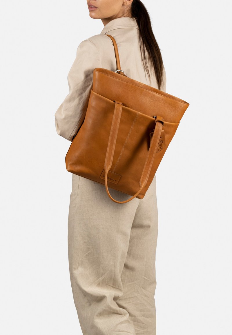 Bolso tote de cuero marrón con dos asas largas, diseño minimalista y un logo visible en la parte frontal. Textura suave y forma estructurada.