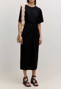 Schwarzes Midikleid mit einem geknoteten Frontdesign, weichem Stoff und kurzen Ärmeln. Kombiniert mit braunen, geschnürten Absatzsandalen und einer beigen geflochtenen Tasche.