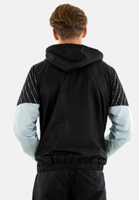 Giacca a vento nera e azzurra chiara con cappuccio, caratterizzata da un pannello nero testurizzato con un motivo sulle maniche. Include polsini ed elastico in vita.